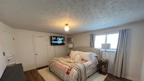 Un dormitorio con cama, televisor montado en la pared, mesita de noche y ventana con cortinas.