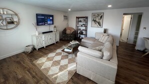 Una sala de estar con un sofá color beige, una mesa de centro, un televisor montado en la pared y una alfombra estampada.