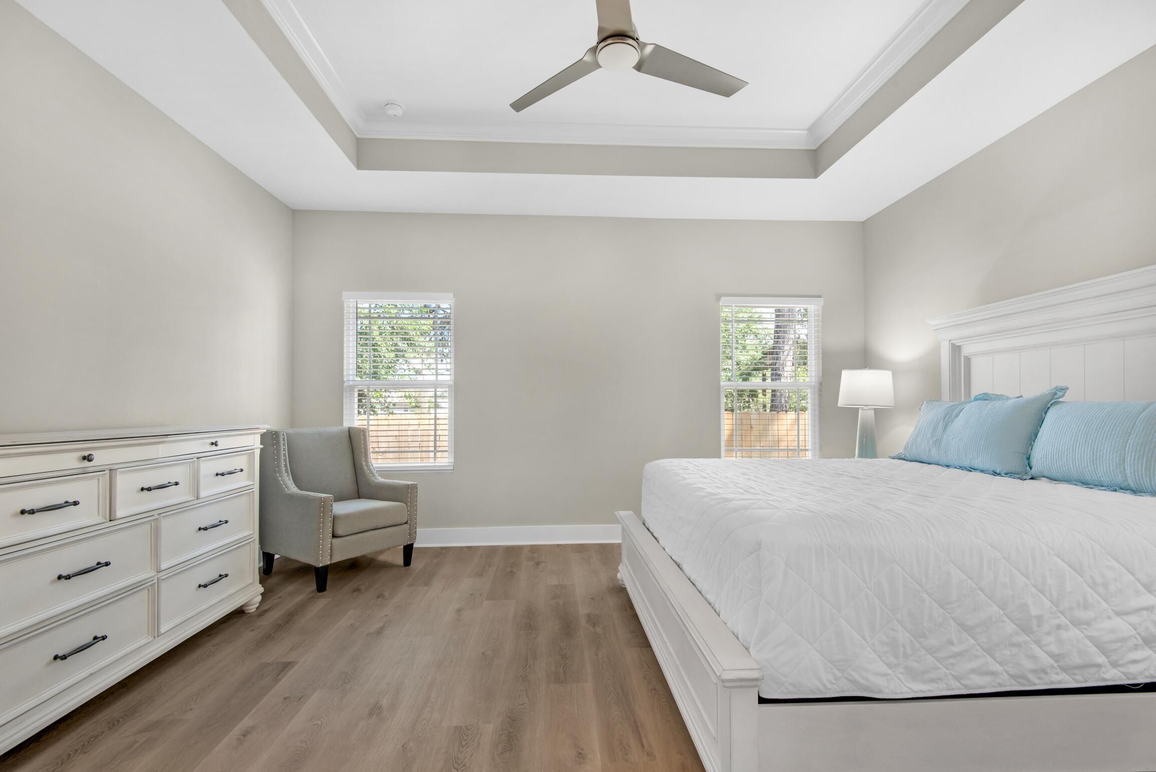 A bedroom with a bed, dresser, chair, ceiling fan, and windows.