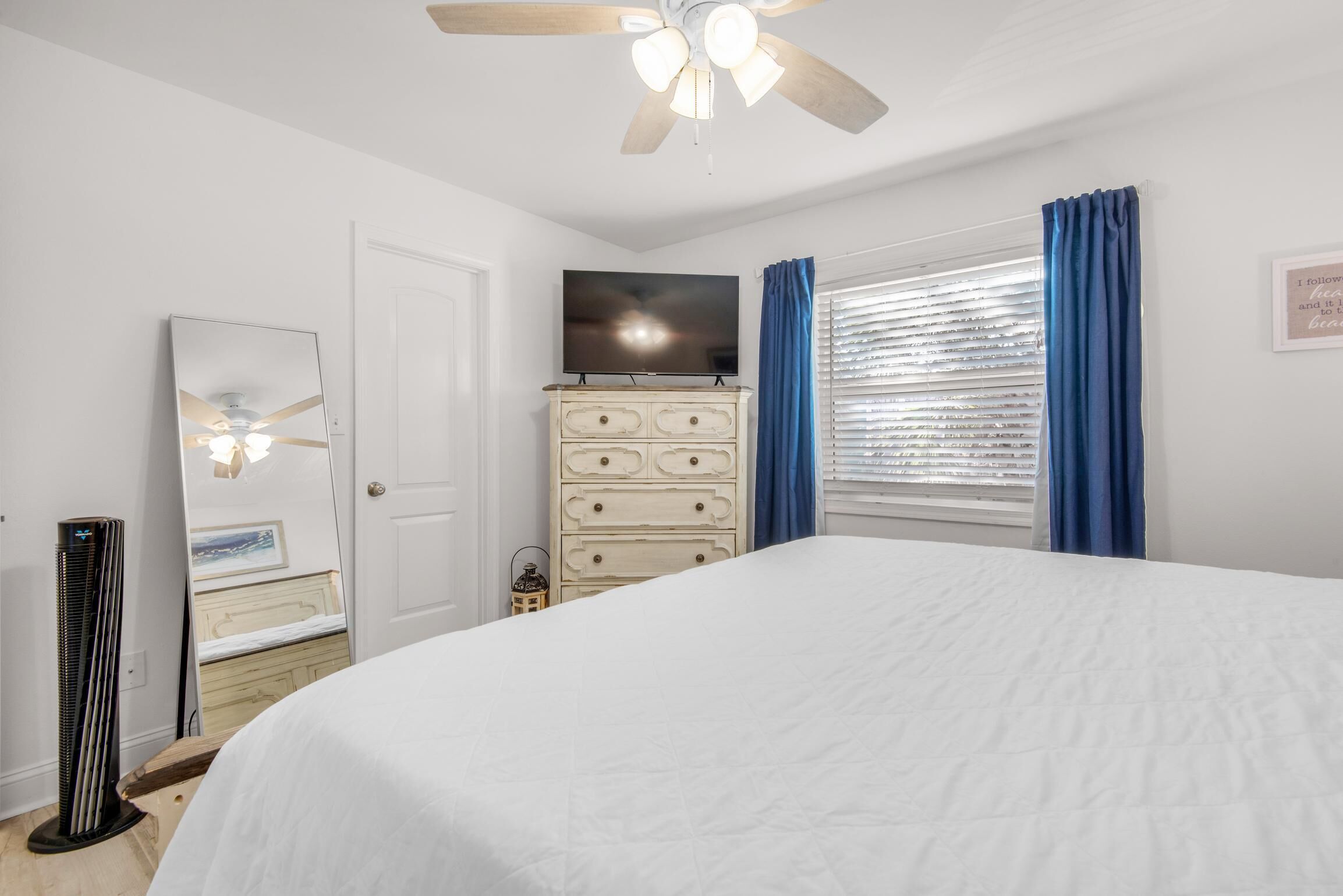 A bedroom with a bed, dresser, ceiling fan, TV, and window with curtains.