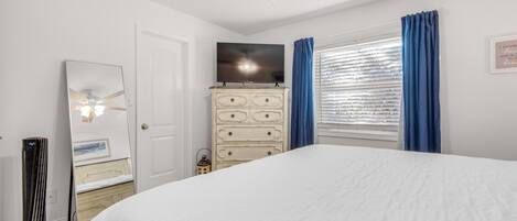 A bedroom with a bed, dresser, ceiling fan, TV, and window with curtains.