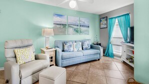 A living room with a sofa, armchair, and ottoman, a ceiling fan, a TV, and artwork on the wall. - Sunset Harbor Villas 313 (Navarre)