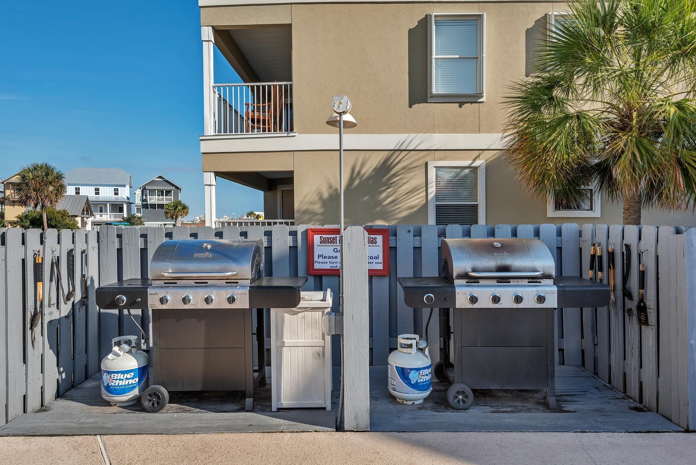 Two barbecue grills with propane tanks, a wooden fence, and a building with balconies in the background.