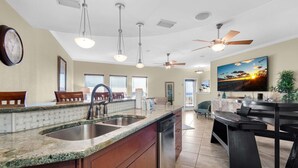 A modern kitchen with granite countertops, stainless steel sink, wooden cabinets, and a dishwasher. There are ceiling fans, pendant lights, and a large window with blinds.