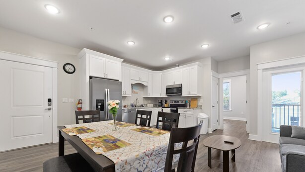A modern kitchen with white cabinets, a dining table with a floral tablecloth, and a small seating area with a gray couch. - Fun in the Sun 2 Bed Apt. (Biloxi)