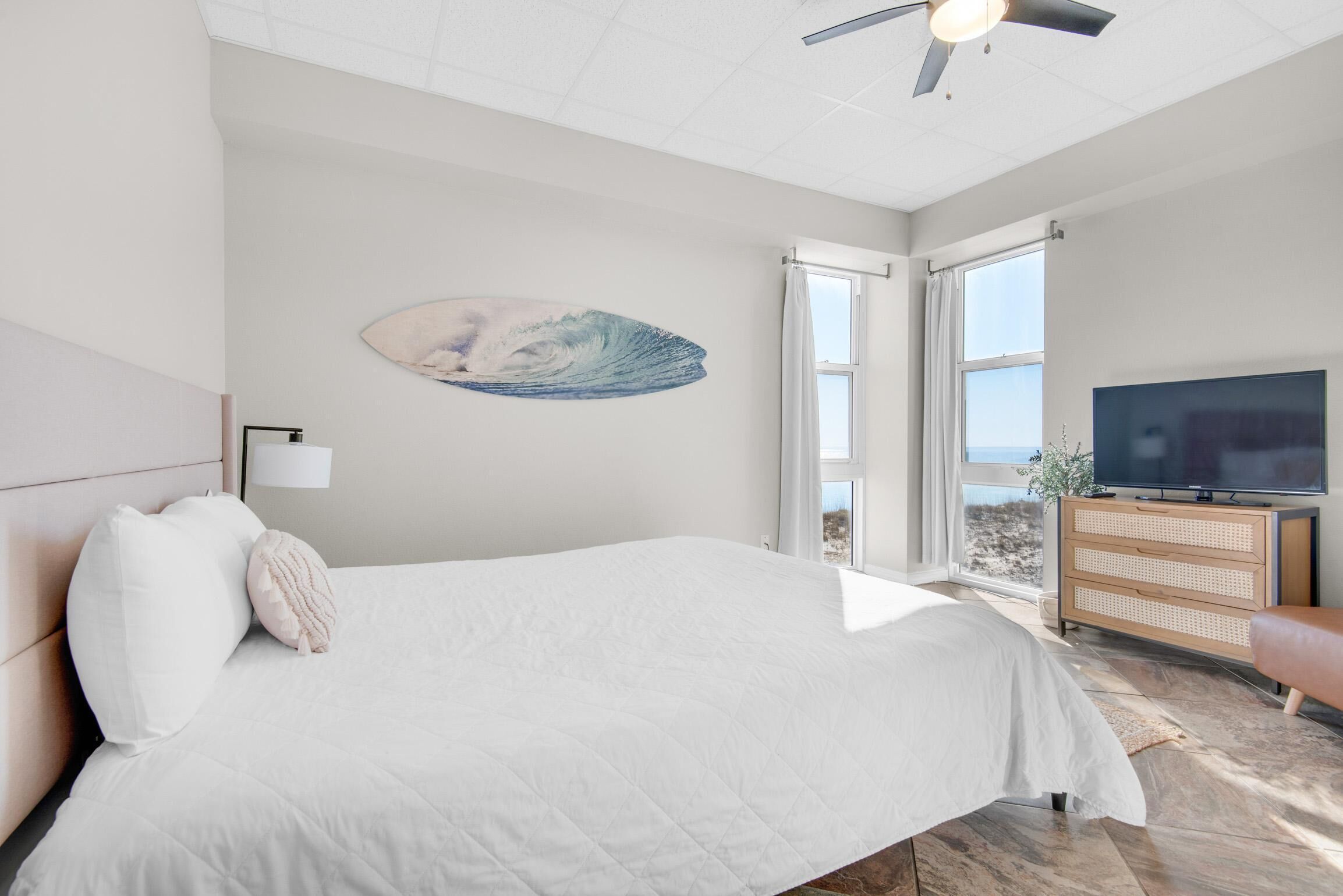 A bedroom with a large bed, a ceiling fan, a TV, and a view of the outdoors.