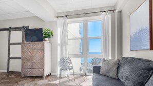 A modern living room with a grey sofa, a wooden cabinet with a TV, a plant, and a view of the sea through large windows. - Sans Souci Unit 209 (Pensacola Beach)
