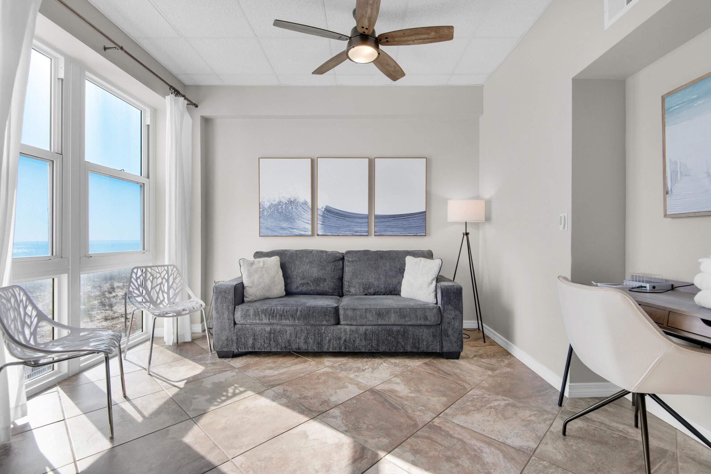 A modern living room with a grey sofa, white chairs, a ceiling fan, and large windows with a view.