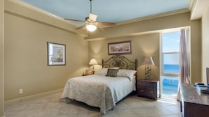 A bedroom with a bed, a nightstand, a lamp, a ceiling fan, a window with a view, and a framed picture on the wall. - Sans Souci Unit 704 (Pensacola Beach)