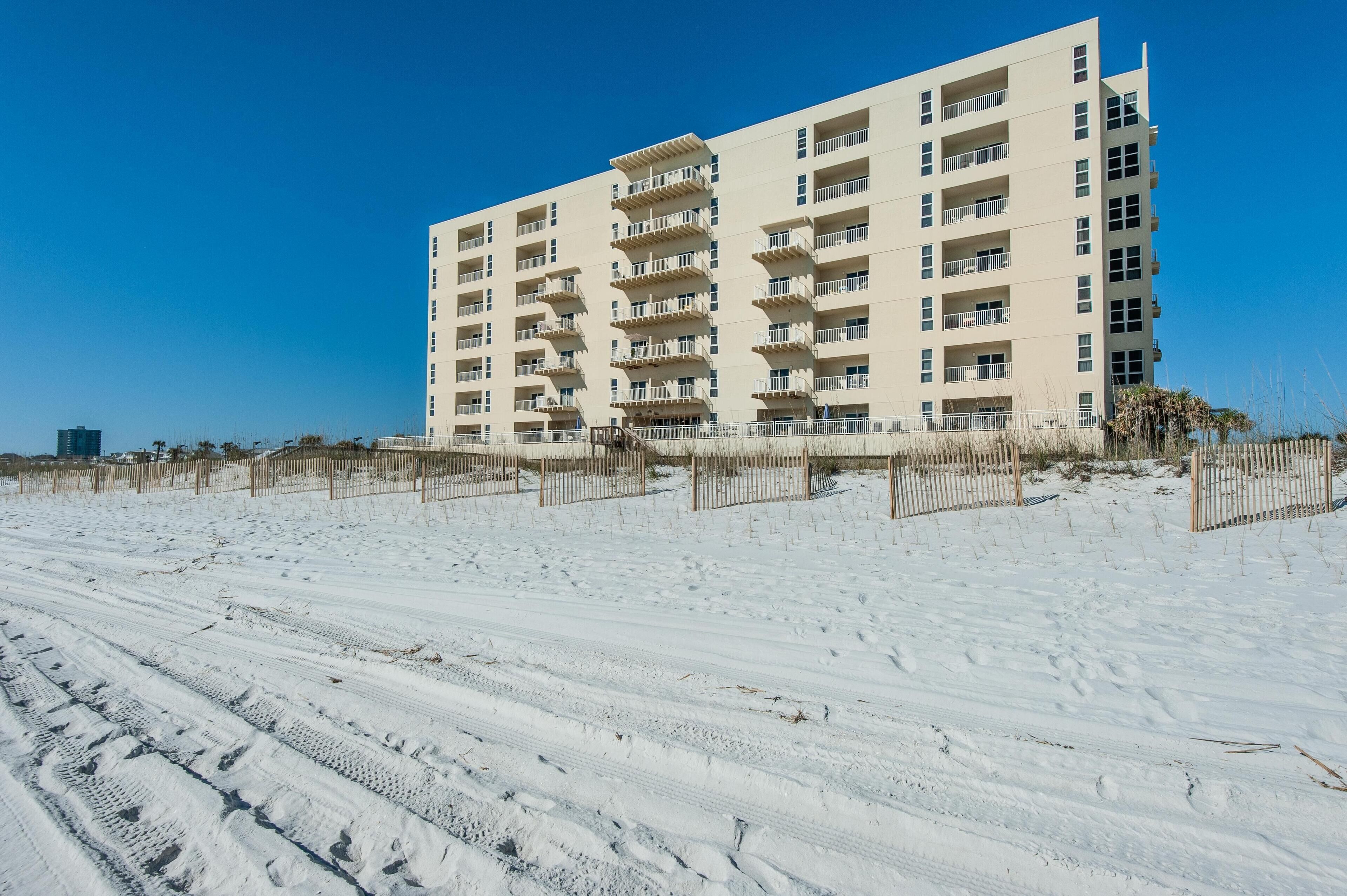 Un hôtel à plusieurs étages doté de balcons, situé sur une plage de sable fin, sous un ciel dégagé.
