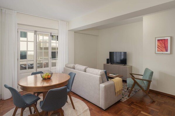 Una sala de estar moderna con sofá, sillas, una mesa de comedor con frutero, televisor y alfombra estampada.