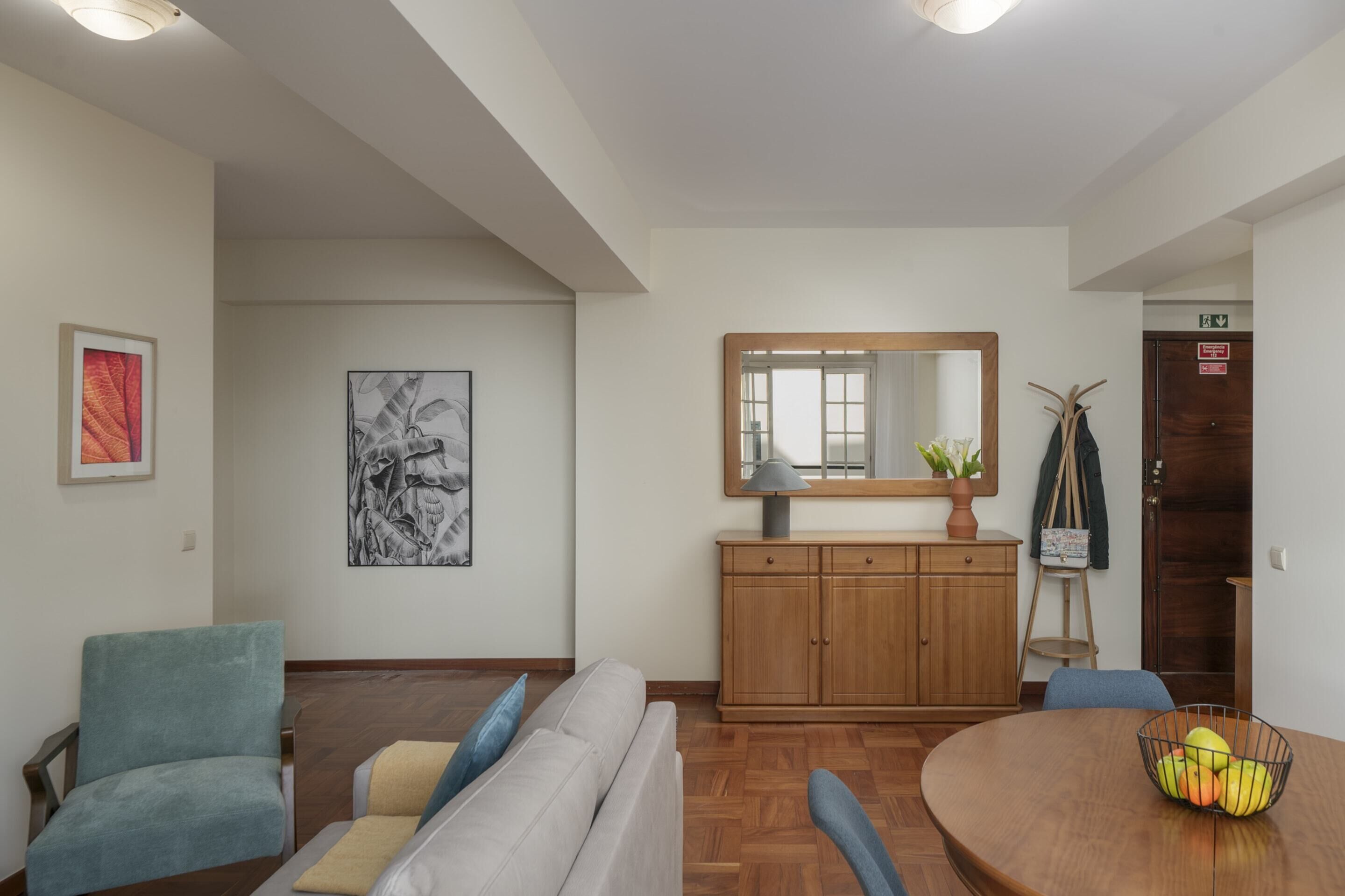 A living room with a sofa, armchairs, a wooden cabinet, and a dining table with a fruit bowl.