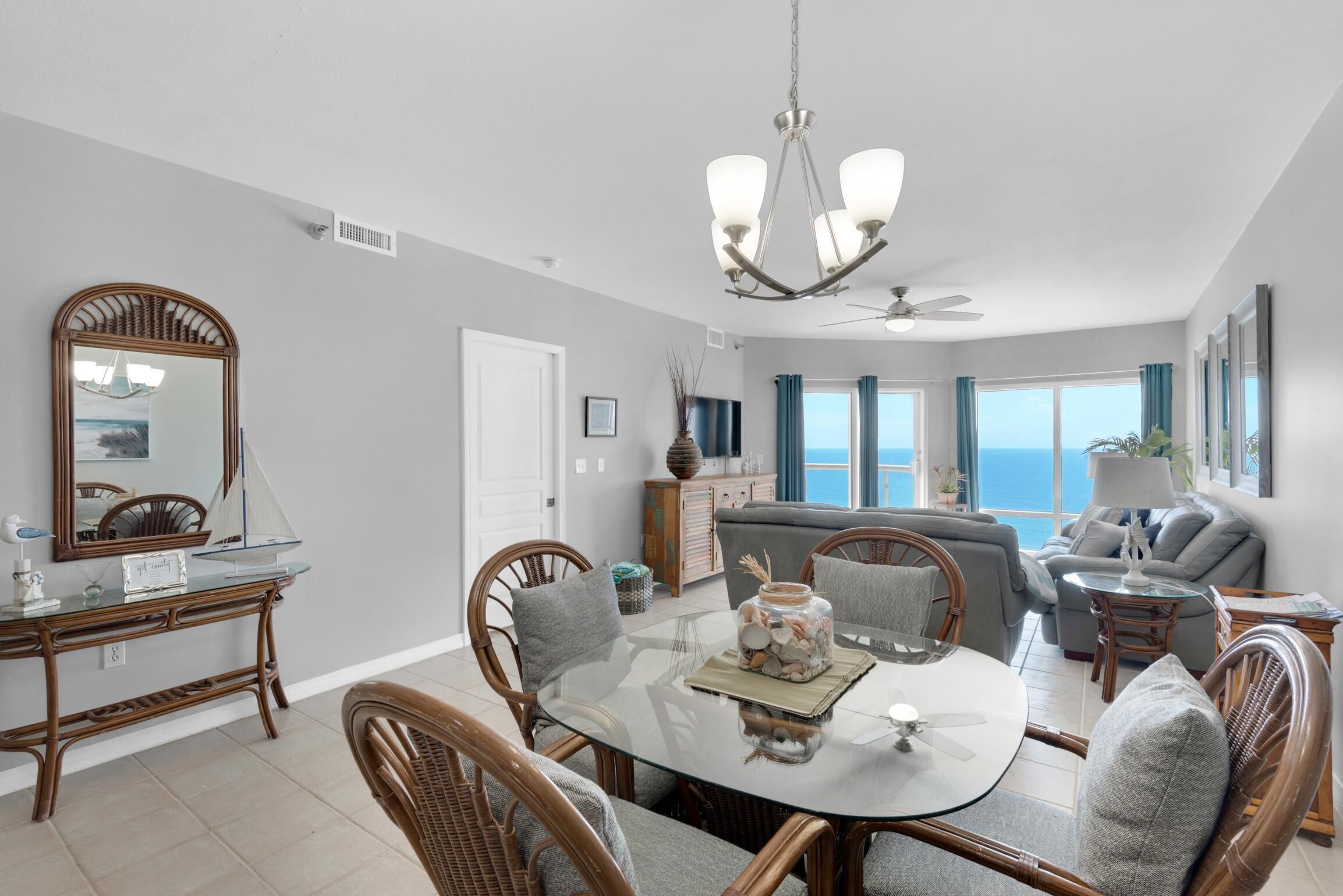 A dining area with a glass table, wicker chairs, a chandelier, and a view of the ocean through large windows.