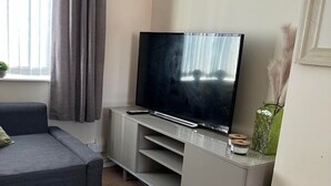 A living room with a grey sofa, a flat-screen TV on a white entertainment unit, a small green vase with dried plants, and a window with grey curtains. - Flat 1,1 bedroom flat sleeps 4 in Abingdon Oxfordshire (Abingdon)