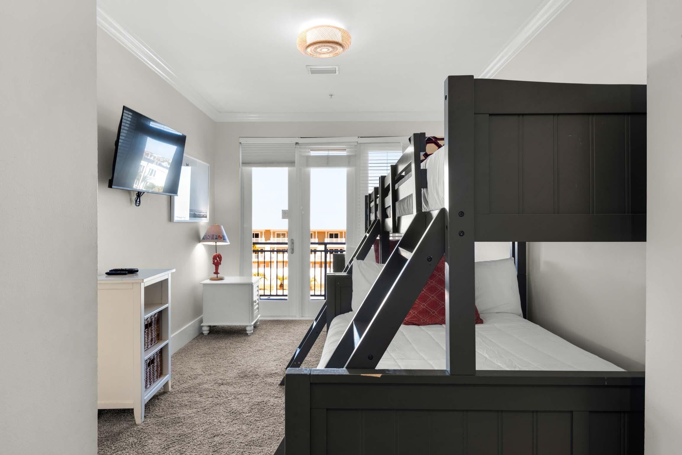 A room with bunk beds, a television, a lamp, a small table, and a view of the outside balcony.
