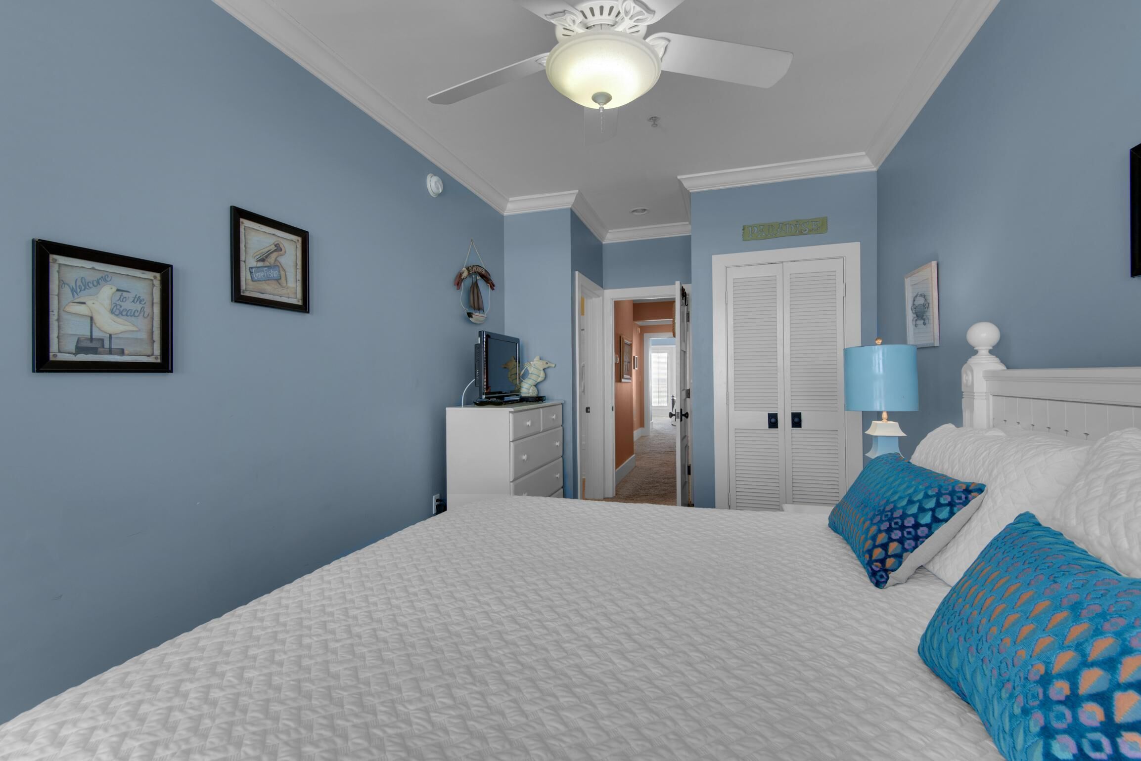 Ein Schlafzimmer mit Bett, Schminktisch, Deckenventilator und einer Tür zu einem weiteren Raum.