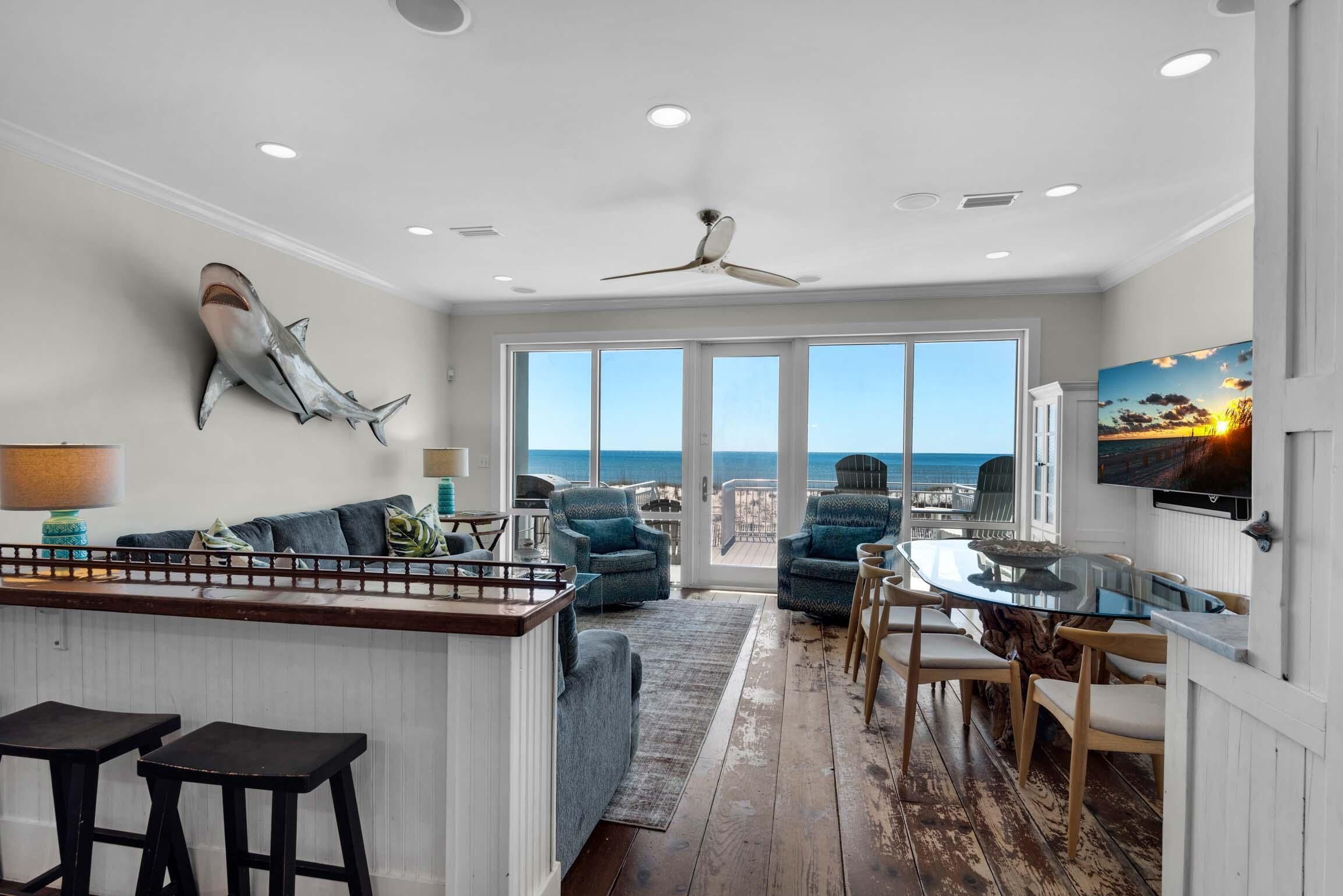 A spacious living area with a large window overlooking the ocean, a ceiling fan, and a bar area with stools.
