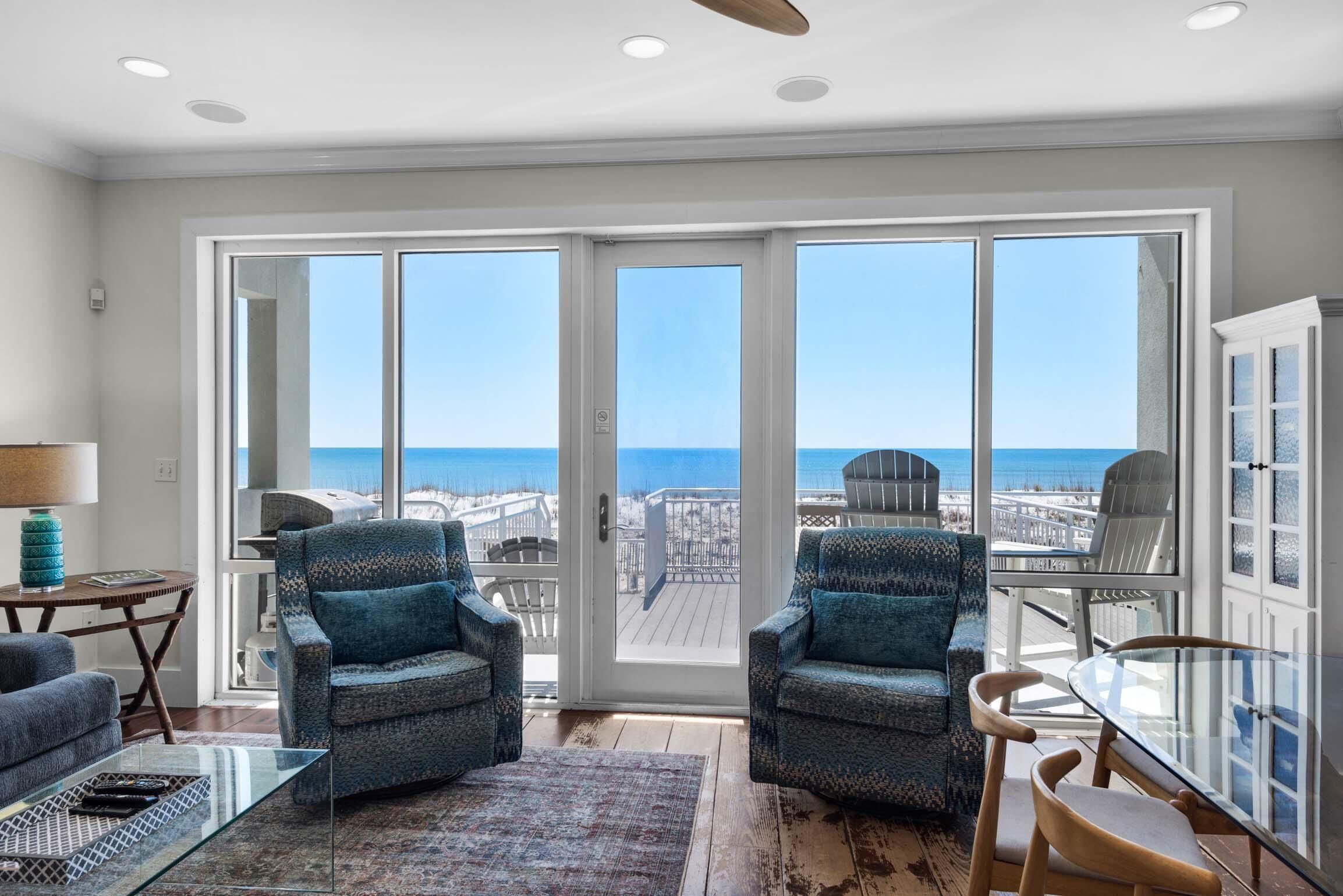A spacious living room with a large sliding glass door leading to a balcony with a view of the ocean. There are two patterned armchairs, a glass coffee table, and a wooden dining table with chairs.