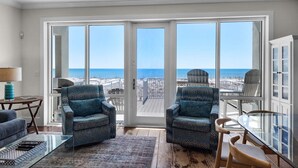 A spacious living room with a large sliding glass door leading to a balcony with a view of the ocean. There are two patterned armchairs, a glass coffee table, and a wooden dining table with chairs. - White Sands 451- Beach Dreams (Pensacola Beach)