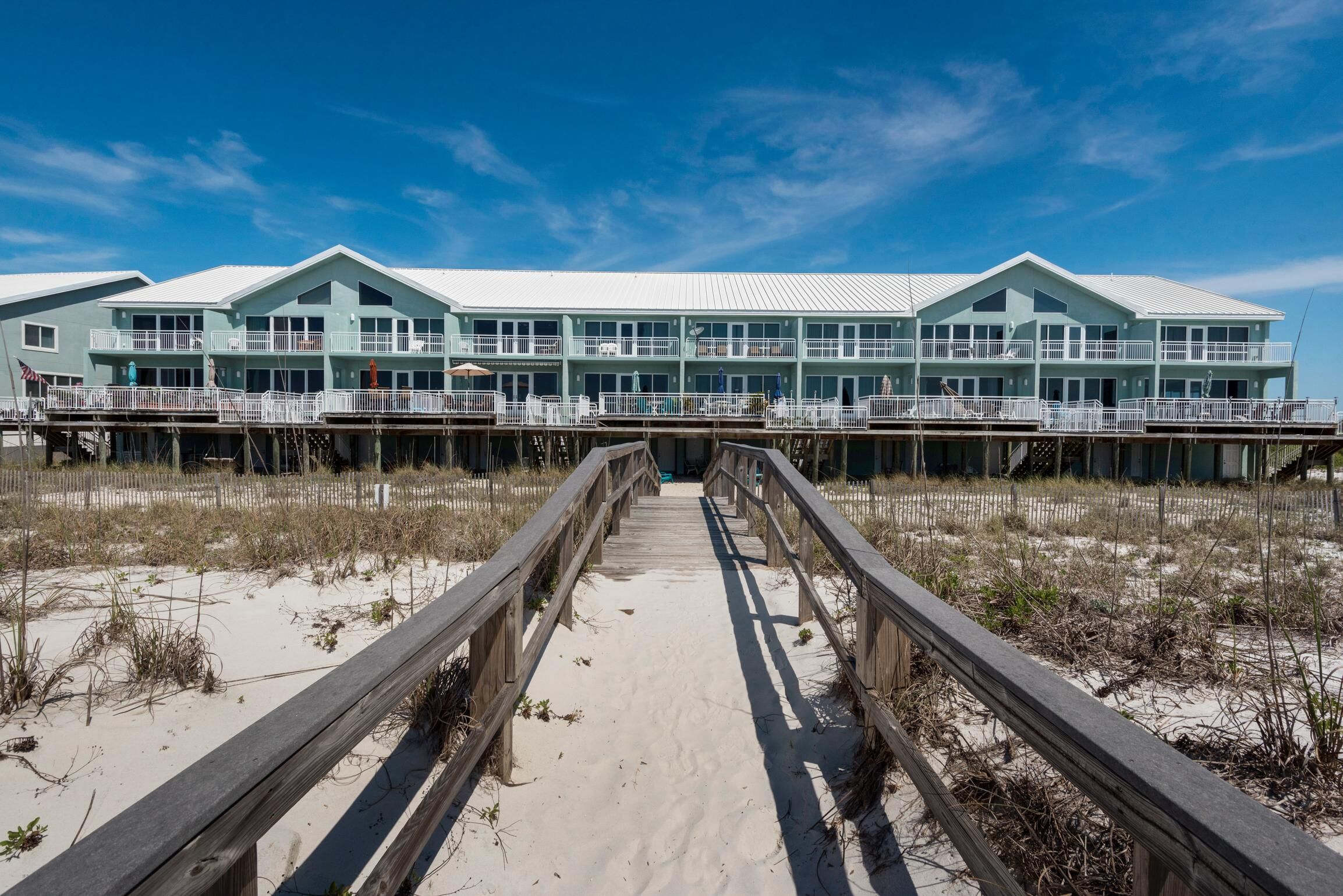 Un hotel frente a la playa con un paseo marítimo de madera que lleva a la entrada, con varios balcones y ventanas amplias.