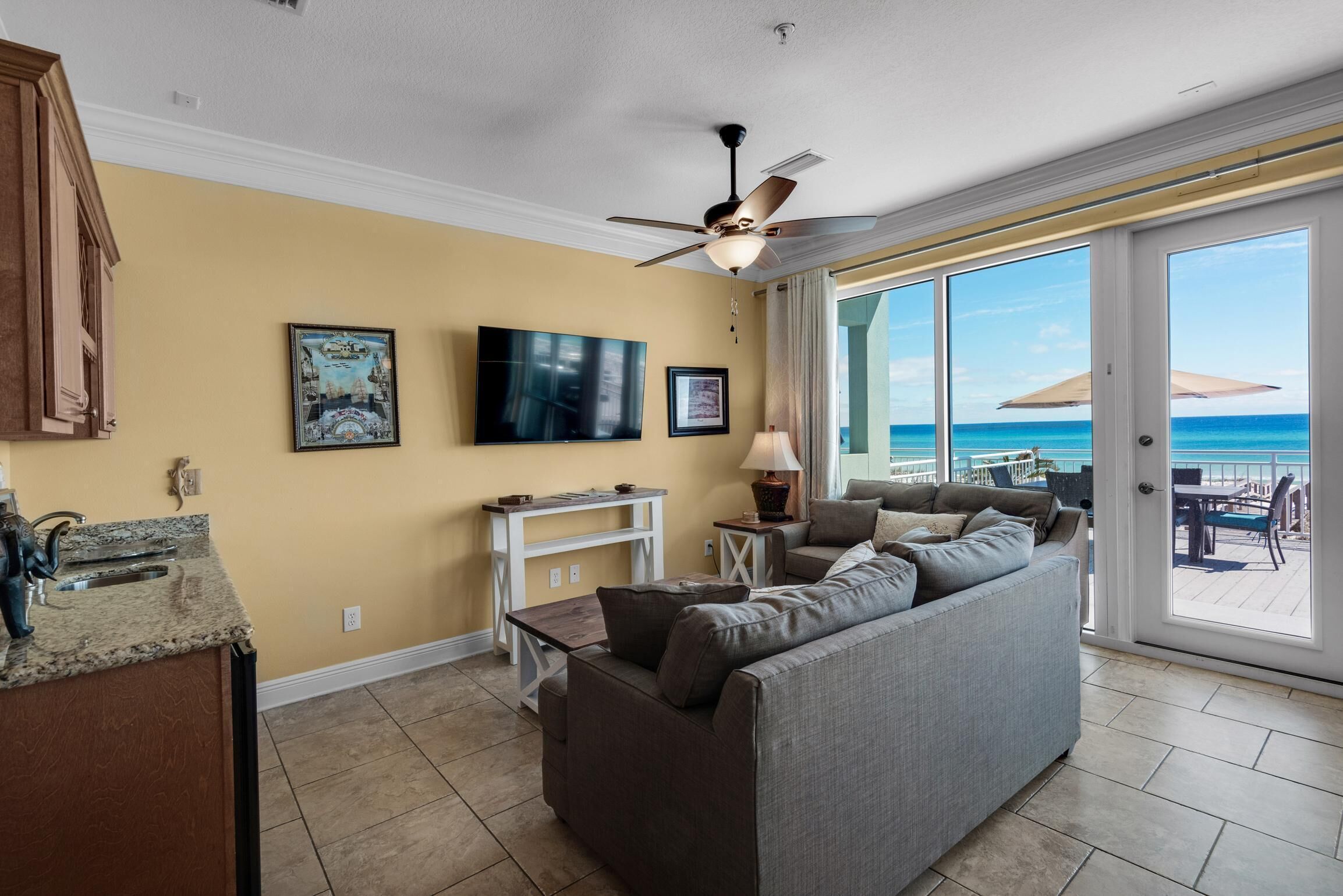 A living room with a gray sofa, a ceiling fan, a TV, and a large window overlooking the ocean.