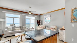 A spacious living area with a large window overlooking the ocean, a kitchen island, and a cozy seating area. - Seahorse Unit R (Pensacola Beach)