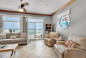 A spacious living room with a large window overlooking the ocean, a ceiling fan, and a view of the beach.