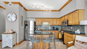 A modern kitchen with wooden cabinets, a stainless steel refrigerator, a microwave, and a granite countertop. There is a clock on the wall and a basket with decorative items on a small table. - Seahorse Unit R (Pensacola Beach)