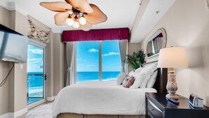 A bedroom with a large bed, a ceiling fan, a TV, a nightstand, a lamp, and a view of the ocean. - Portofino Island Resort P5-1003 (Pensacola Beach)