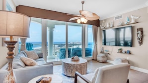 A spacious living room with a large window, a ceiling fan, a round coffee table, and a sofa. - Portofino Island Resort P5-1003 (Pensacola Beach)