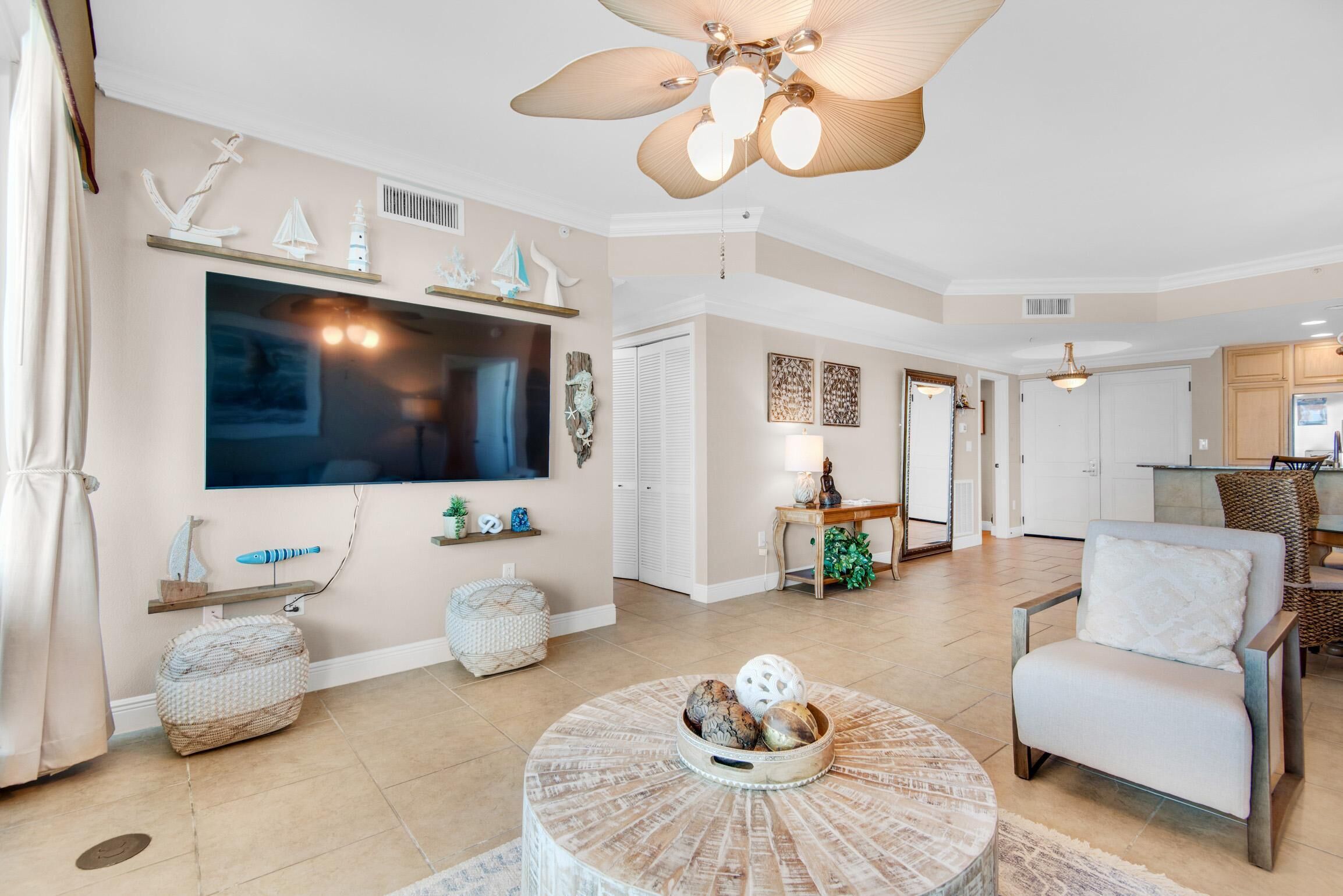 A spacious living room with a large TV, a round coffee table with decorative items, a ceiling fan, and a view into the kitchen area.