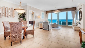 A spacious living room with a dining area, large windows offering a view of the ocean, and a ceiling fan. - Portofino Island Resort P5-1003 (Pensacola Beach)