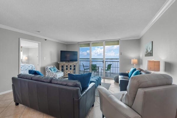 A modern living room with a grey sofa, a beige armchair, and a television on a cabinet. A balcony with a view of the sky and clouds is visible through large windows.