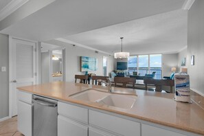 A modern kitchen with a large island, a dishwasher, and a sink. There is a dining area with a bench and a view of the outdoors.