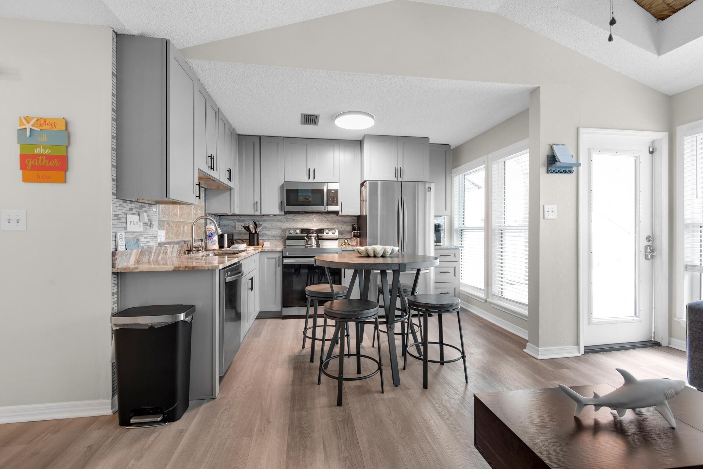 A modern kitchen with grey cabinets, a stainless steel refrigerator, a microwave, and a wooden countertop. There are four stools around a central table.