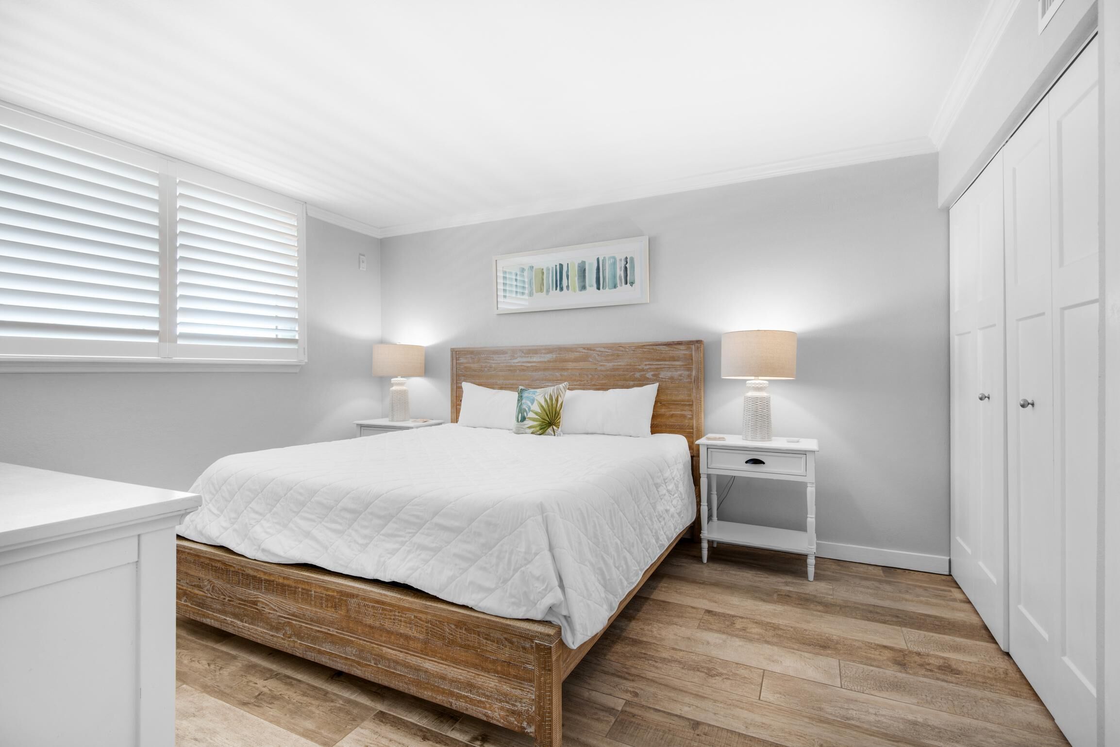 Un dormitorio con cama de madera, ropa de cama blanca, dos mesitas de noche con lámparas, un armario blanco y una ventana con persianas.