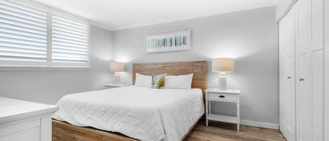 Un dormitorio con cama de madera, ropa de cama blanca, dos mesitas de noche con lámparas, un armario blanco y una ventana con persianas.