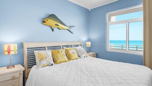 A bedroom with a bed, a nightstand, a lamp, a fish wall art, and a window with a view of the sea. - Seahorse Unit H (Pensacola Beach)