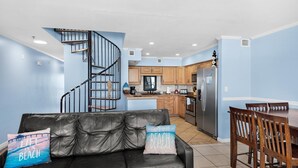 A modern living room with a black leather sofa, wooden dining table, and chairs. A spiral staircase leads to the kitchen area with wooden cabinets and a refrigerator. - Seahorse Unit H (Pensacola Beach)