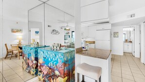 A modern kitchen with white cabinets, a blue island with a coral mural, and a bar area with a white countertop. - Sundunes 112 (Navarre)