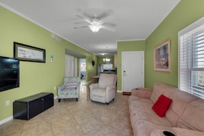 A living room with a beige sofa, a patterned armchair, a black entertainment unit, and a ceiling fan. - Villas on the Gulf Unit J1 (Pensacola Beach)