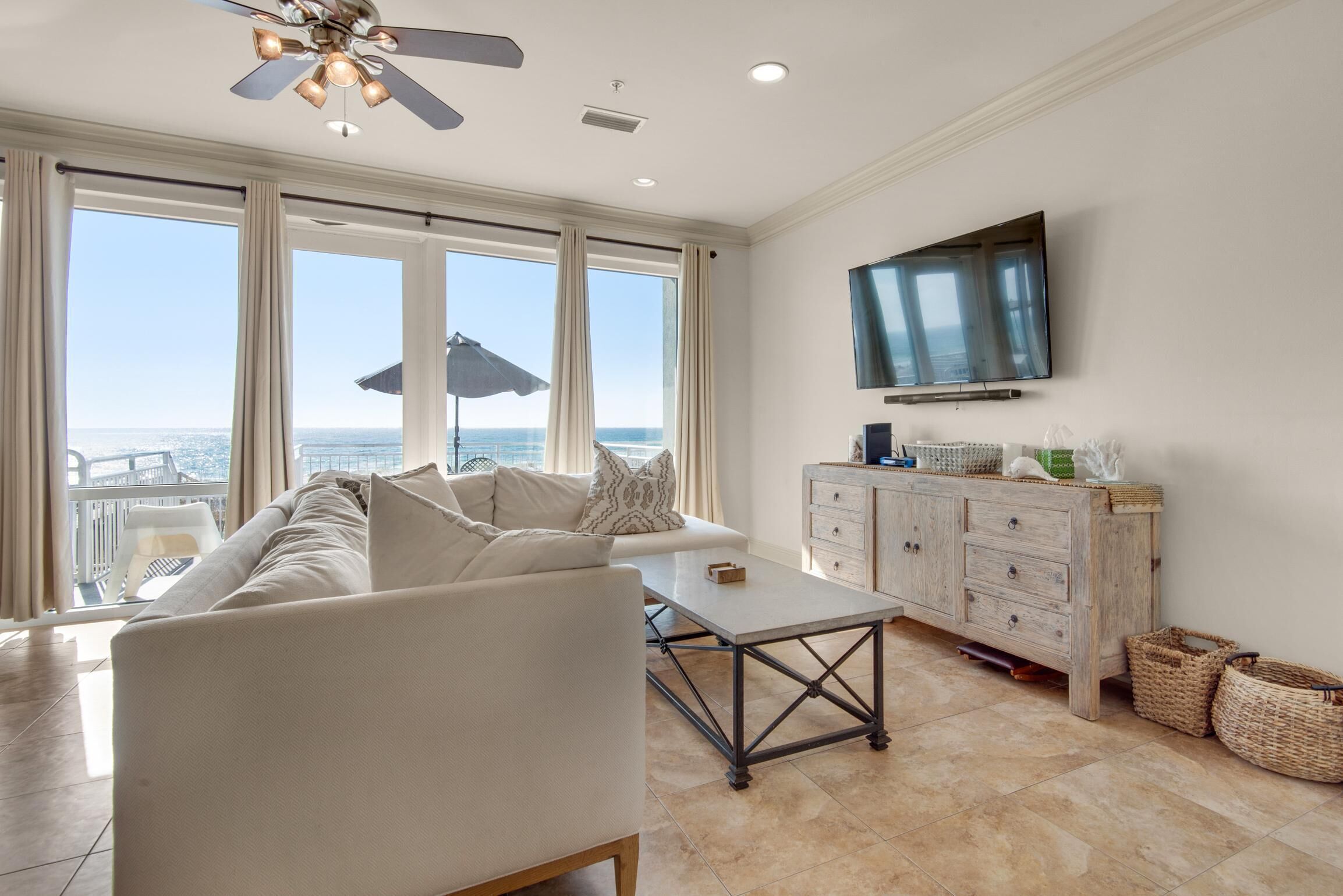 A spacious living room with a large window offering a view of the ocean, a ceiling fan, a flat-screen TV, and a wooden cabinet with baskets.