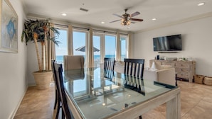 A spacious dining area with a glass table and chairs, a ceiling fan, a TV, and a view of the ocean. - White Sands 447 (Pensacola Beach)