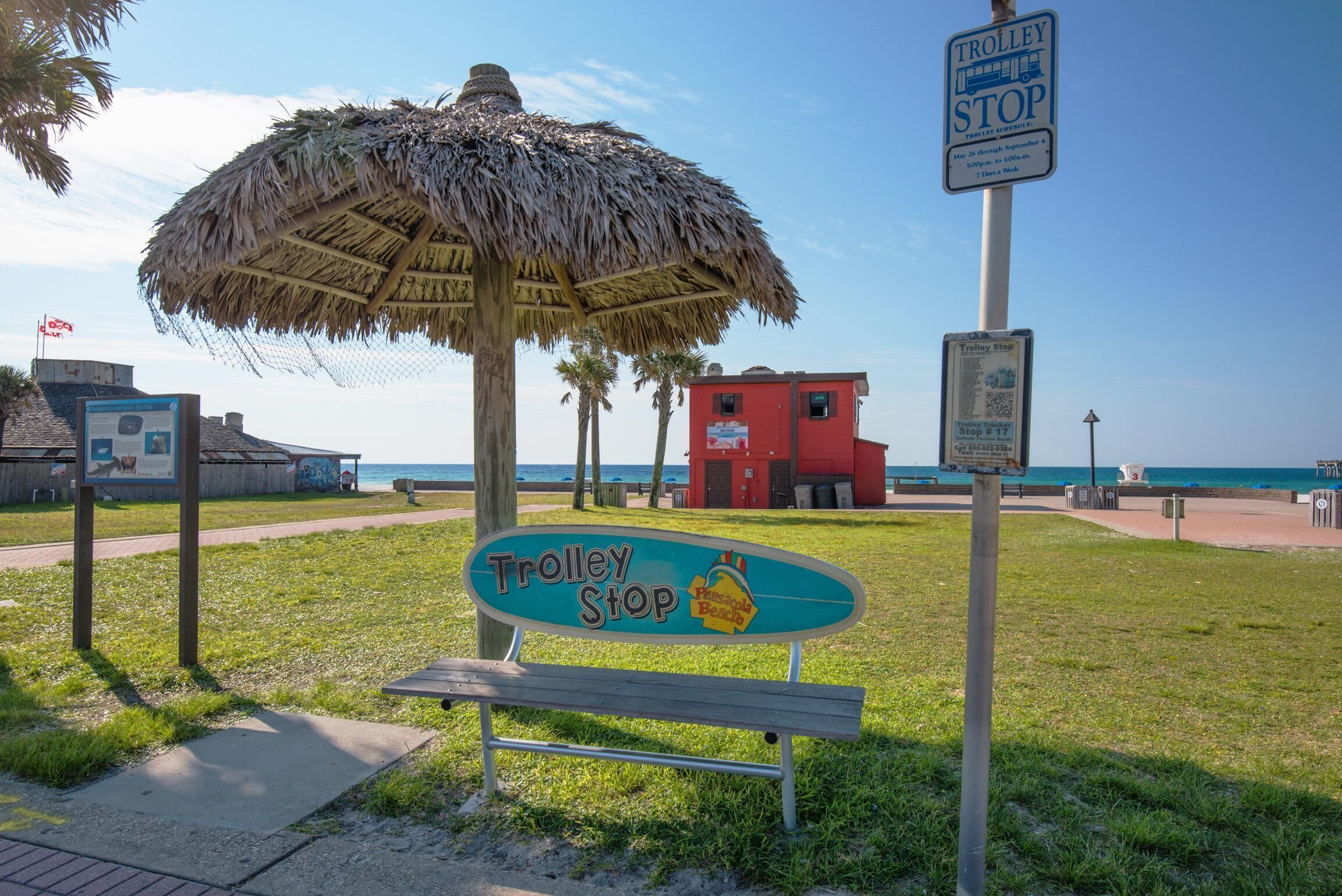 A beachside trolley stop with a thatched umbrella, a red building, and a bench.