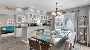 A modern kitchen with white cabinets, a wooden dining table with blue placemats, and a large window with a view of the city. - Regency Cabanas E1 (Pensacola Beach)