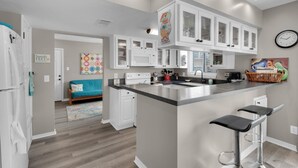 A modern kitchen with white cabinets, a central island with bar stools, a microwave, and a clock on the wall. - Regency Cabanas E1 (Pensacola Beach)