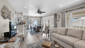 A spacious living area with a sofa, coffee table, and a TV on a stand. There is a dining area with a table and chairs, and a ceiling fan. - Regency Cabanas E1 (Pensacola Beach)
