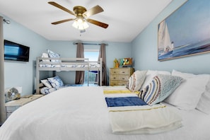 A bedroom with a bunk bed, a bed with a white comforter, a ceiling fan, a dresser, a television, and a painting of a sailboat. - Boardwalk B2 (Pensacola Beach)