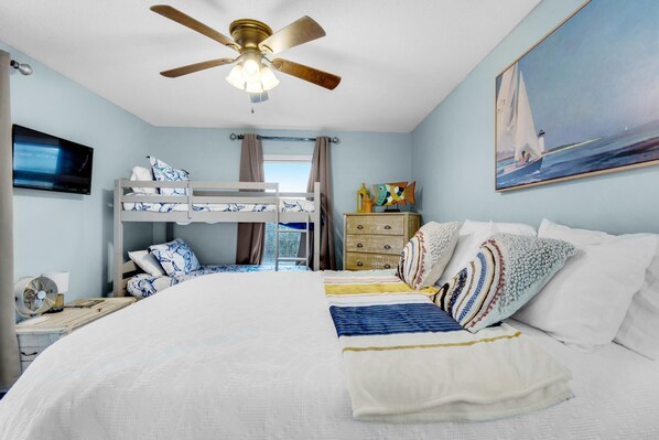 A bedroom with a bunk bed, a bed with a white comforter, a ceiling fan, a dresser, a television, and a painting of a sailboat.