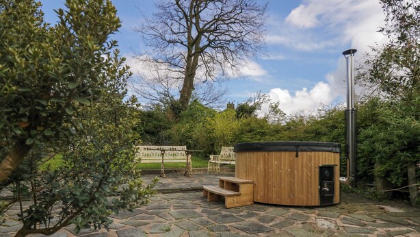 A hot tub in a garden with stone flooring, surrounded by greenery and a bench. - Eden Vale (Broughton in Furness)
